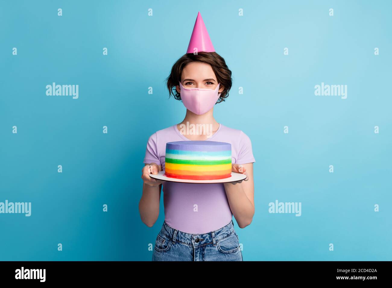 Portrait of positive cheerful girl in medical mask celebrate covid quarantine birthday hold cake wear cone lilac violet denim jeans isolated over blue Stock Photo