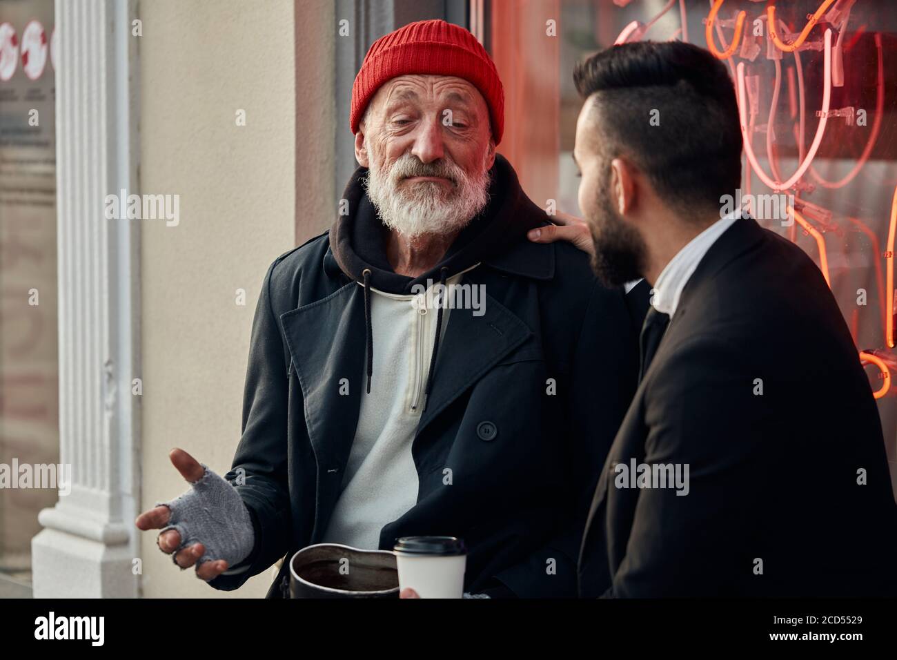 Compassionate man in black tuxedo sit listening to beggar male's story about his life in street, talking Stock Photo