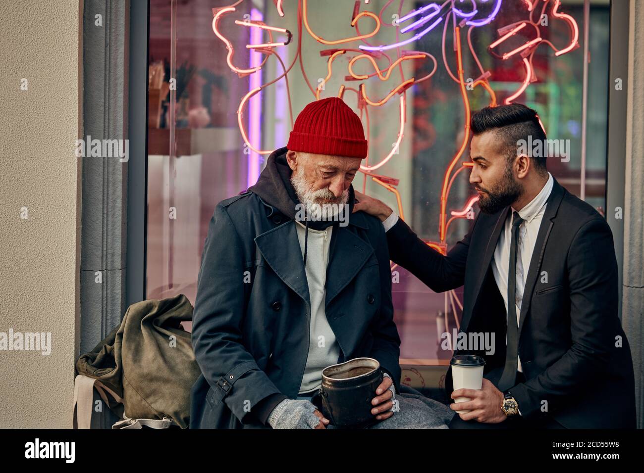 Businessman in tuxedo and homeless person in dirty clothes sitting and talking in the street, look at each other. Poverty, help Stock Photo