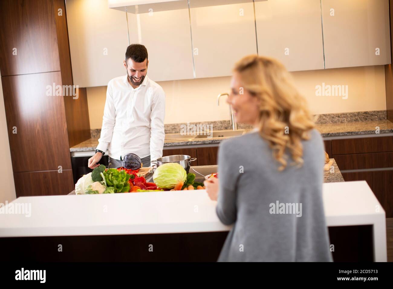 Young woman drinking coffee at the kitchen table while man preparing food Stock Photo