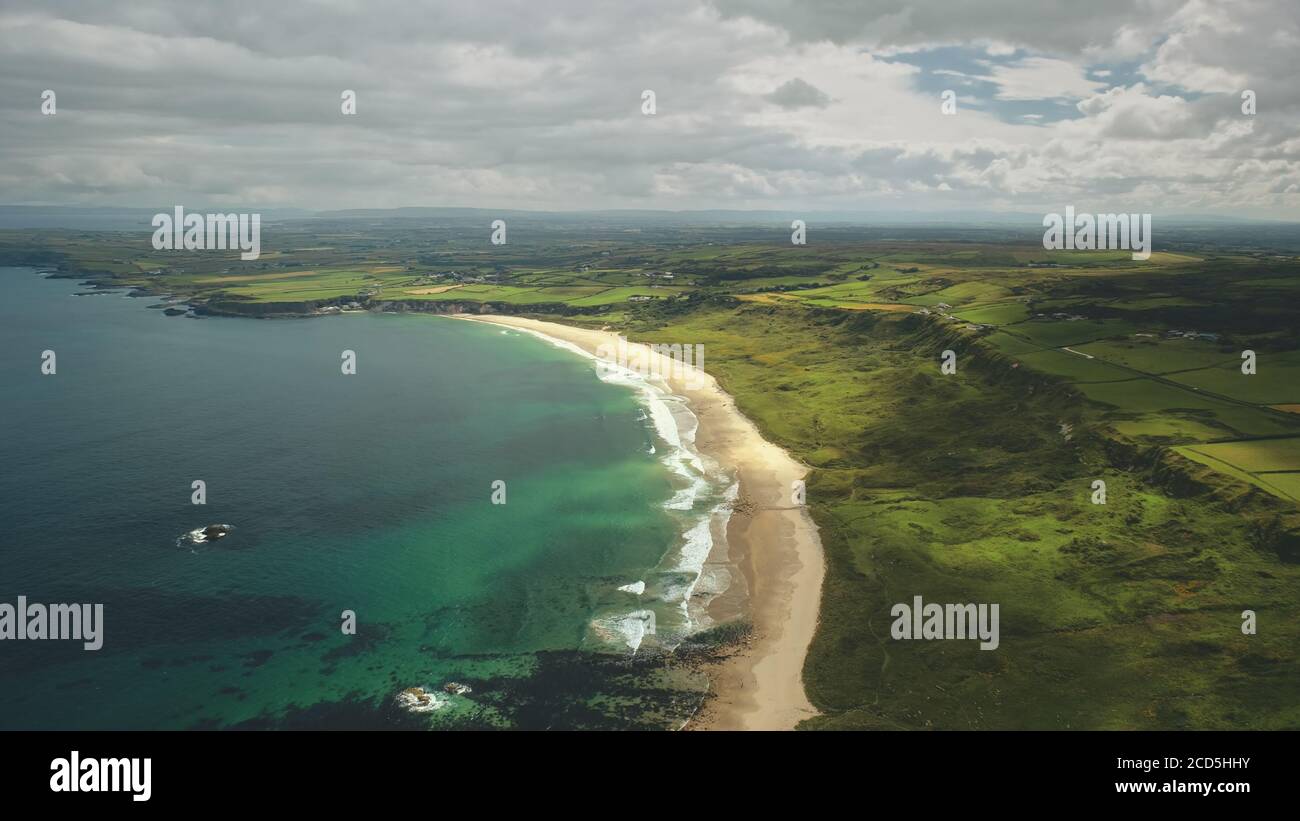 Aerial view sandy coastline, green grass meadows. Waves crashing out of shore and come back to ocean. Atlantic bay serene scenery in dusk summer daytime cinematic shot Stock Photo