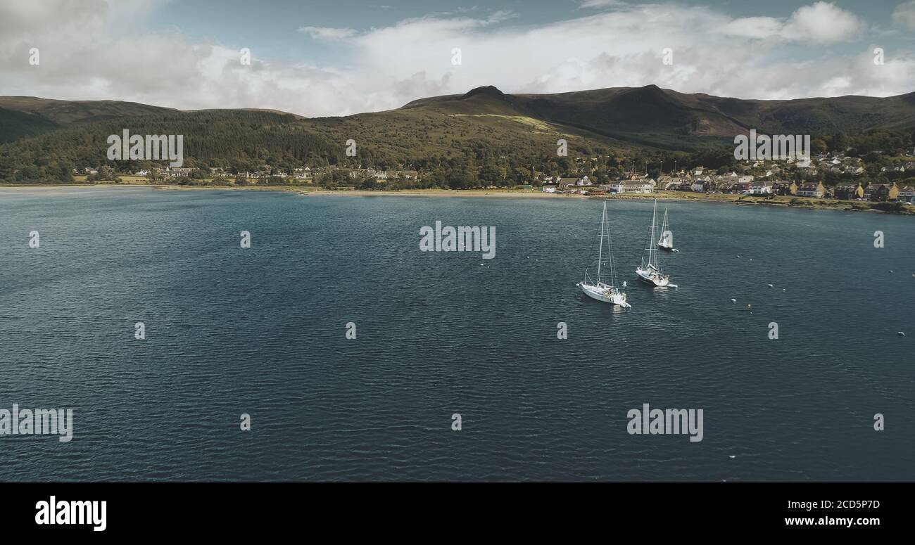 Atlantic ocean gulf, sailboats aerial zooming shot in Brodick Bay. Scottish landscape of port town. Houses, cottages, resort at shore of gulf against greenery lands and mountains view Stock Photo