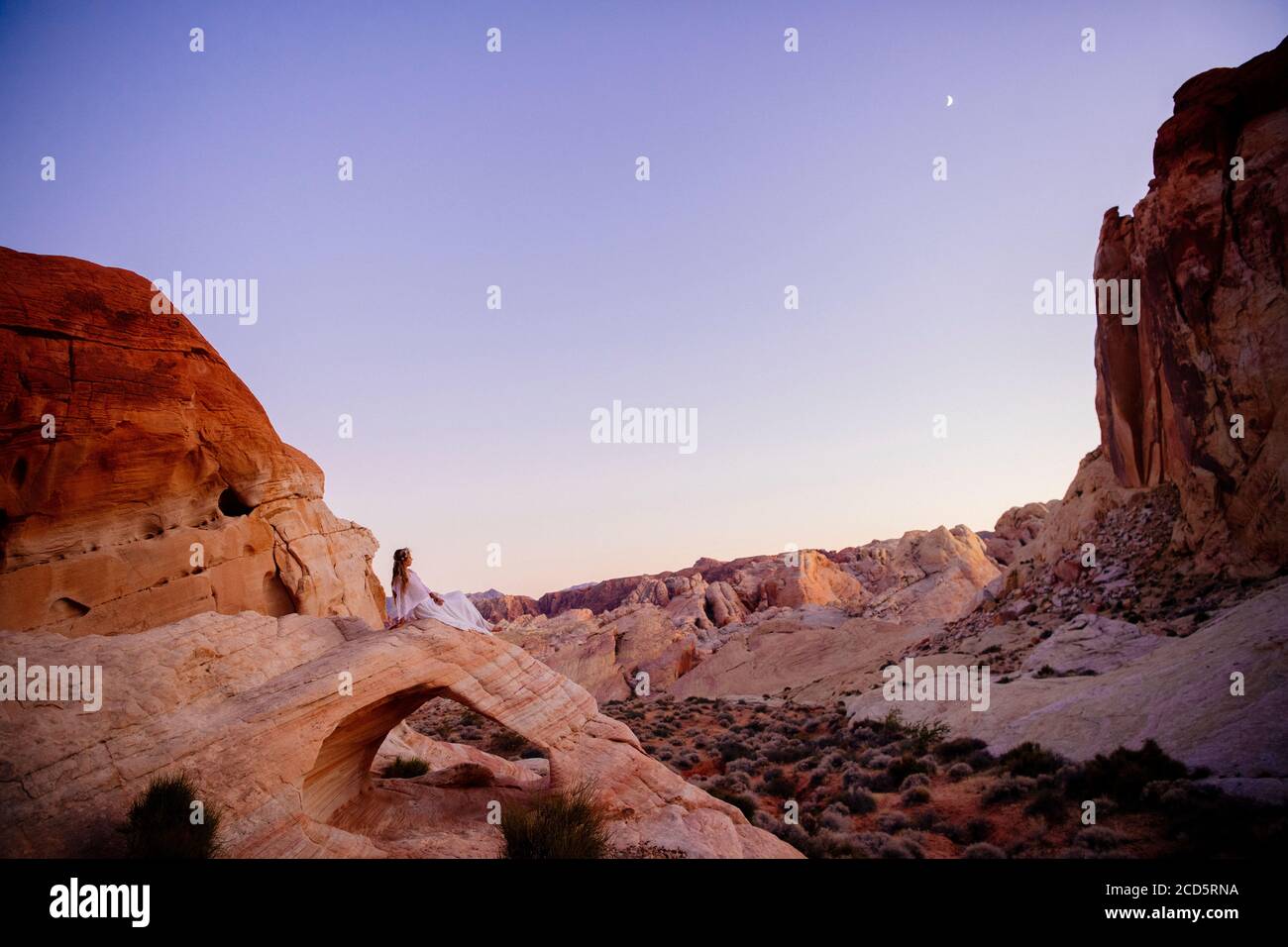 Nymph-like woman in white sitting on Aztec Sandstone natural arch,  State Park, Mohave Desert, Overton, Nevada, USA Stock Photo