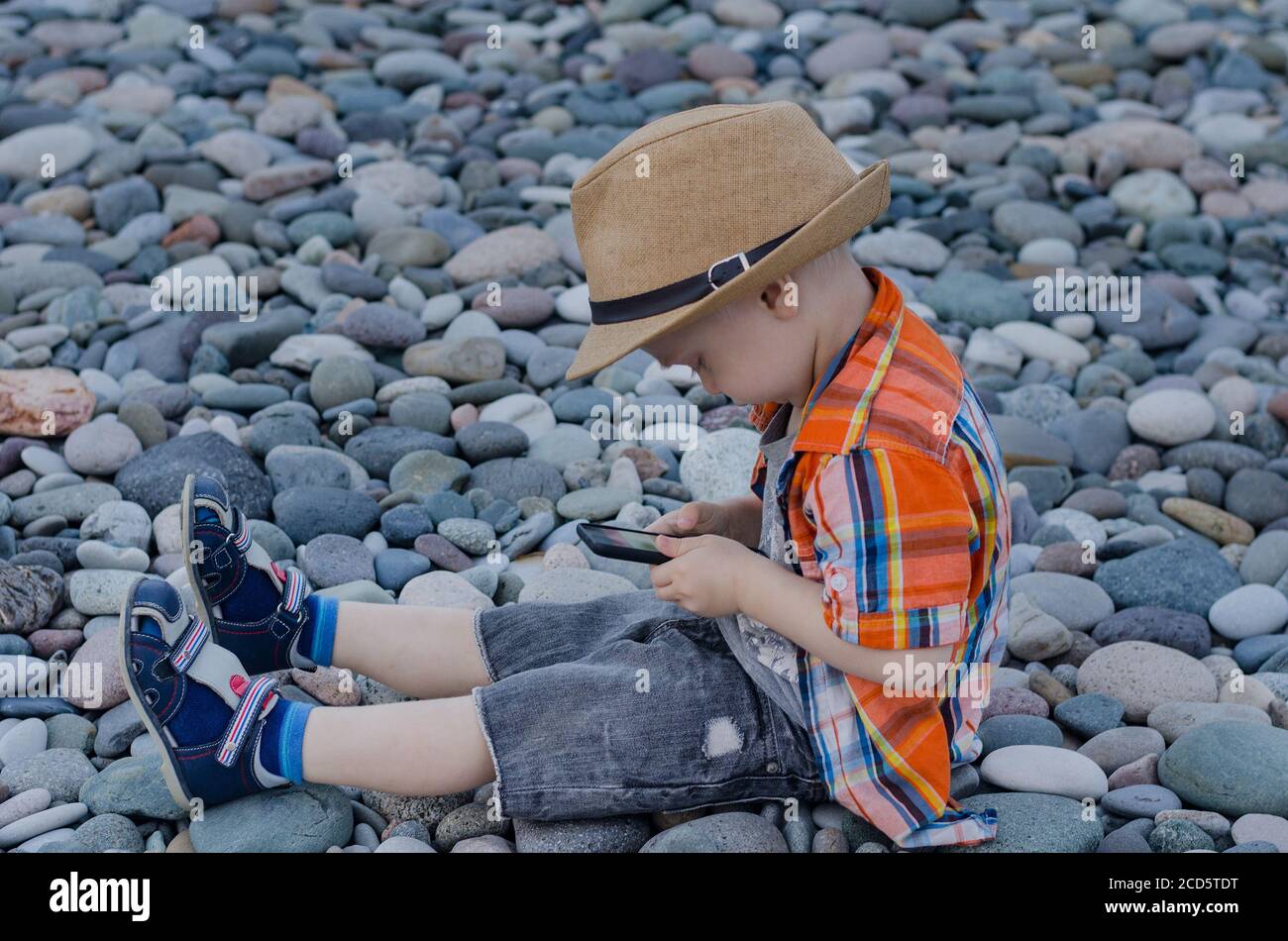 Little boy in straw hat sitting on the pebbles and looking at the phone  Stock Photo - Alamy