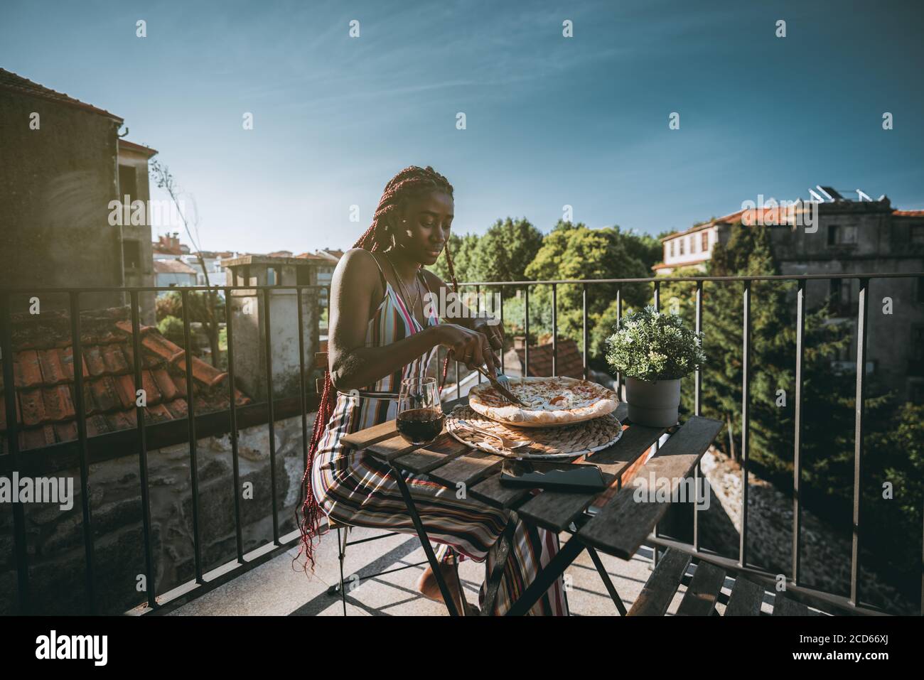 An adorable young black woman is having lunch and cutting into pieces delicious pizza with cheese and bacon while sitting on a balcony Stock Photo