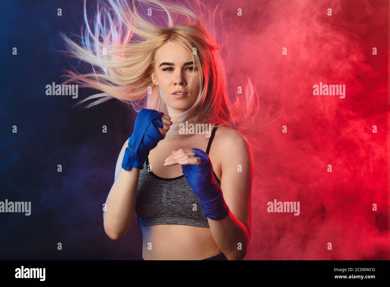 Blonde female athlete is exercising in defense and attacks on dark smoky background. Woman in tank-top and boxing bandages is having cardio boxing wor Stock Photo