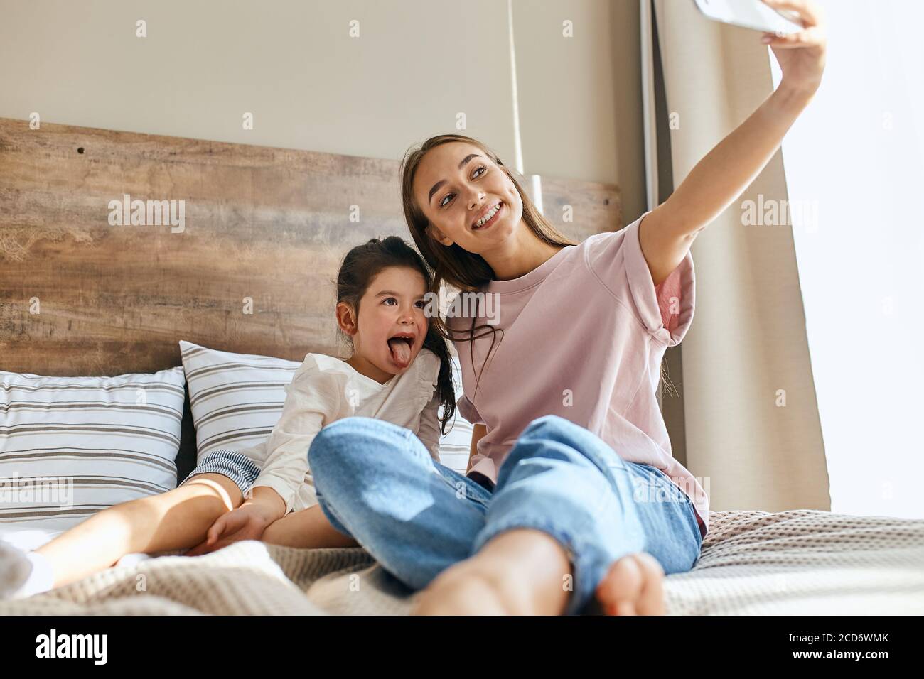 Bottom shot of charming beautiful mother and little child casually dressed, sitting on bed with joyful faces, hugging taking selfie using cell phone, Stock Photo