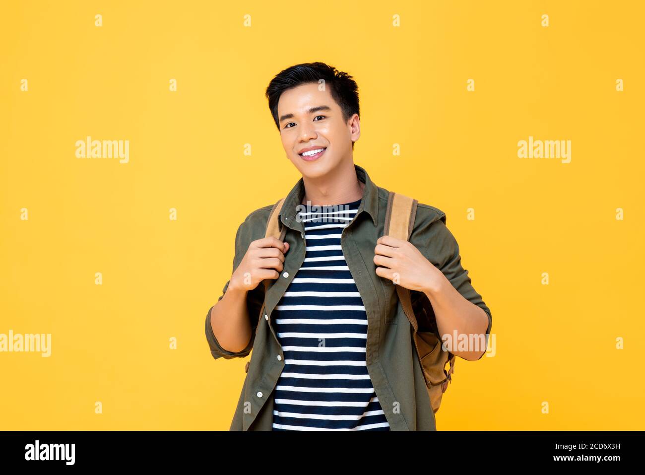 Portrait of smiling young male Asian tourist carrying backpack ready to travel in isolated studio yellow background Stock Photo