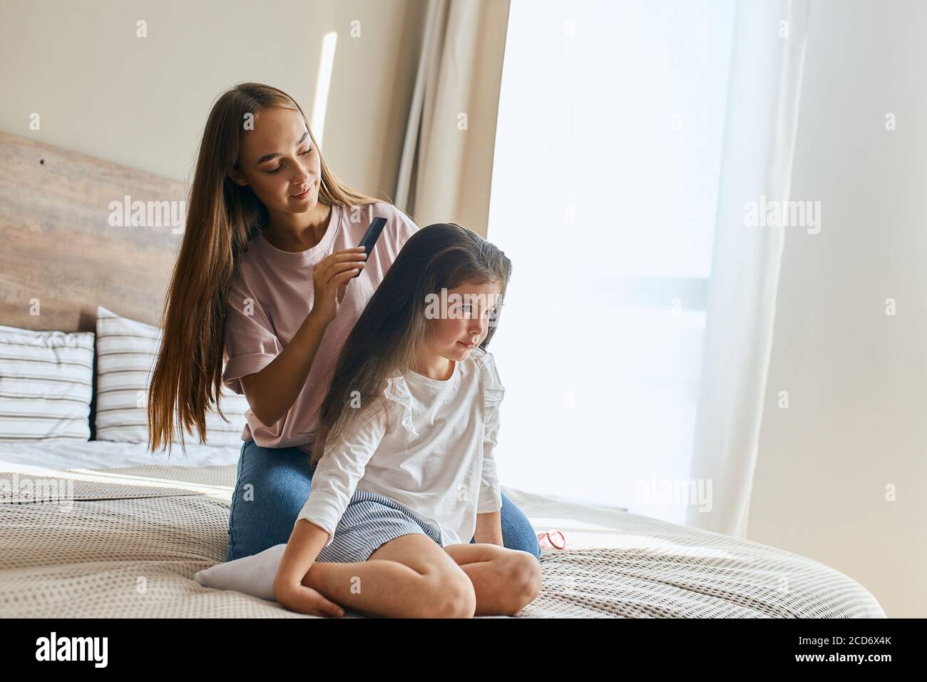 Beautiful charming girl in home clothes, sitting on knees on big bed together with mom, looking aside with bright eyes and calm face, posing against b Stock Photo