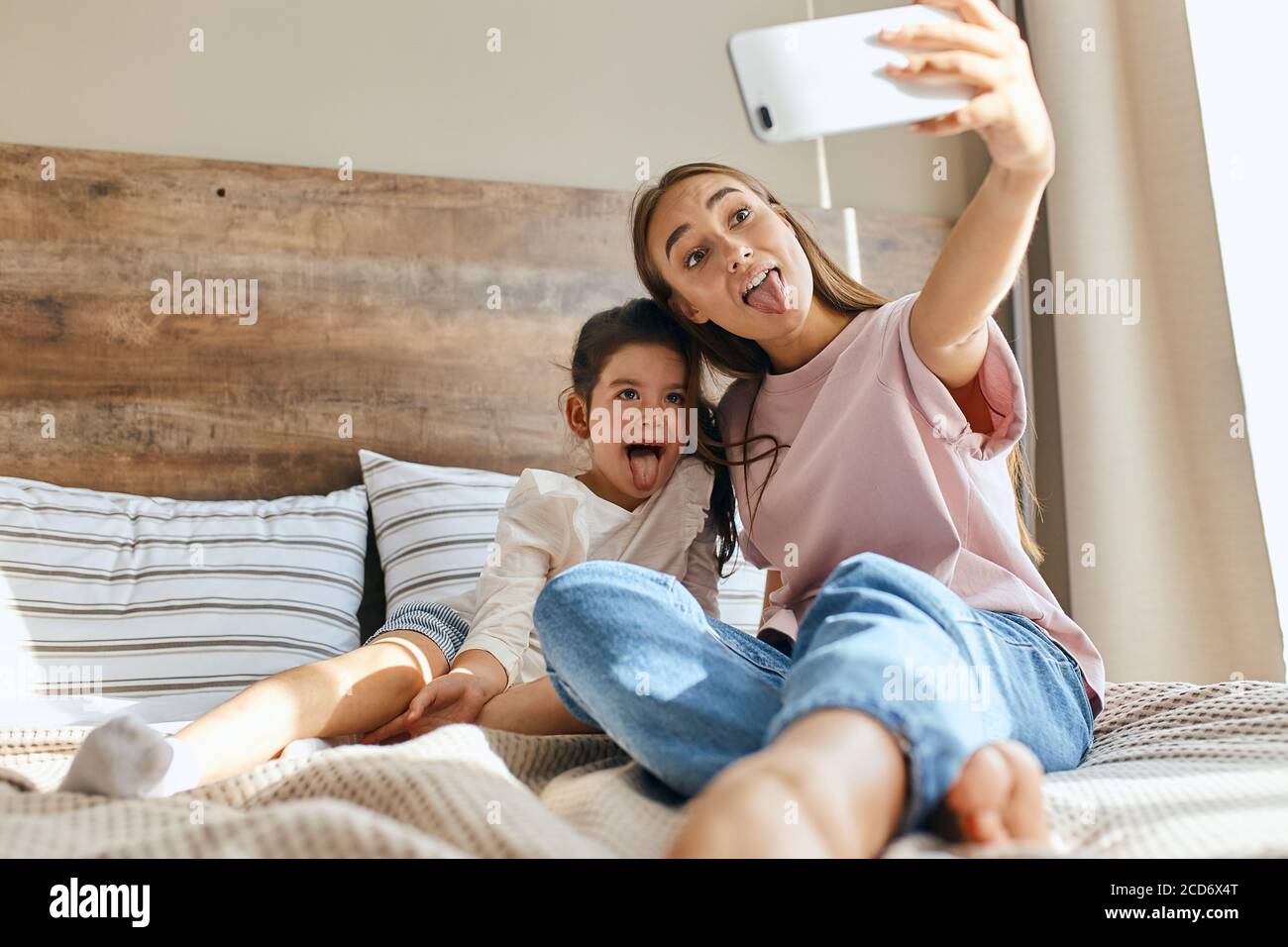 Two beautiful girls having fun in bedroom, attractive woman takes photo using cell phone, showing tongue full of joy, background of small pillows, mor Stock Photo