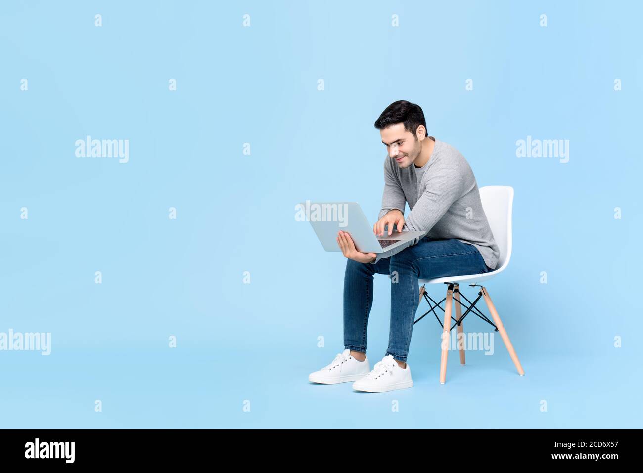 Portrait of smiling young handsome Caucasian man sitting while working on his laptop in isolated studio blue background Stock Photo