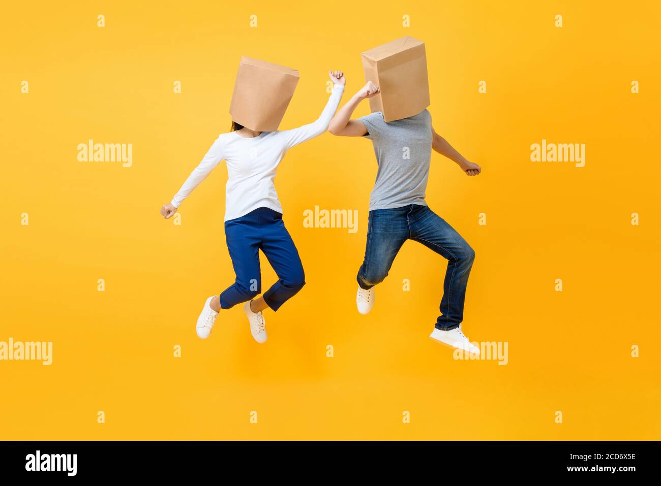 Fun portrait of faceless anonymous couple covering heads with paper bags jumping in mid-air isolated on yellow studio background Stock Photo