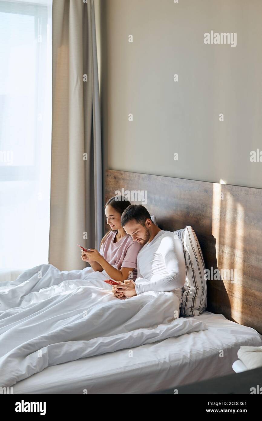 Happy pleasant man and woman spending time together in hotel room, lying in bed under white blanket, staring sell phones screens, ignoring live commun Stock Photo