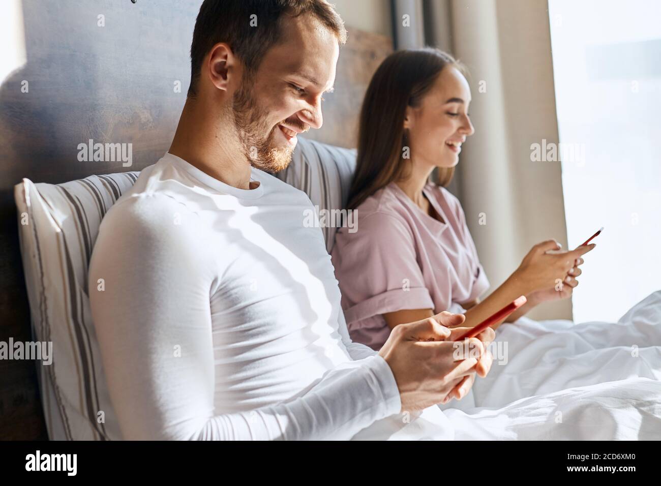 Pleasant young wife and husband chatting with parents using cell phones, getting good news, smiling broadly, looking aside with happy faces, in bedroo Stock Photo
