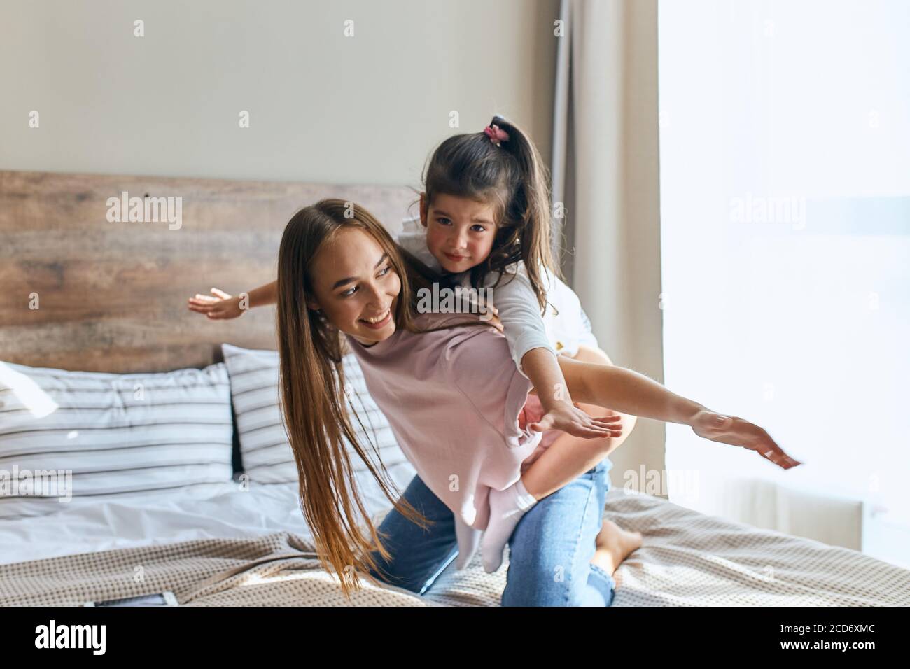 Attractive charming little girl on a piggy back ride with lovely young mom, looking at camera with calm face, woman with long hair playing with child Stock Photo
