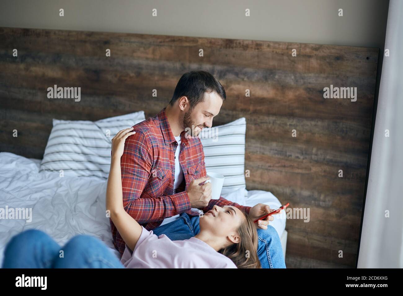 romantic couple dressed in casual outfit, resting in bedroom, pretty girl lying on bed, putting head on knees husband, looking up with happy expressio Stock Photo