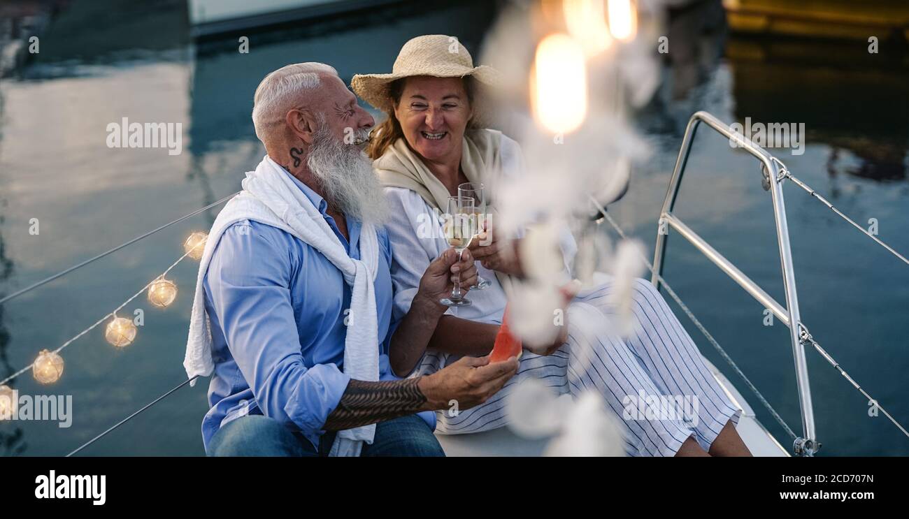 Senior couple toasting champagne and eating fruits on sailboat vacation - Happy elderly people having fun celebrating wedding anniversary on boat trip Stock Photo