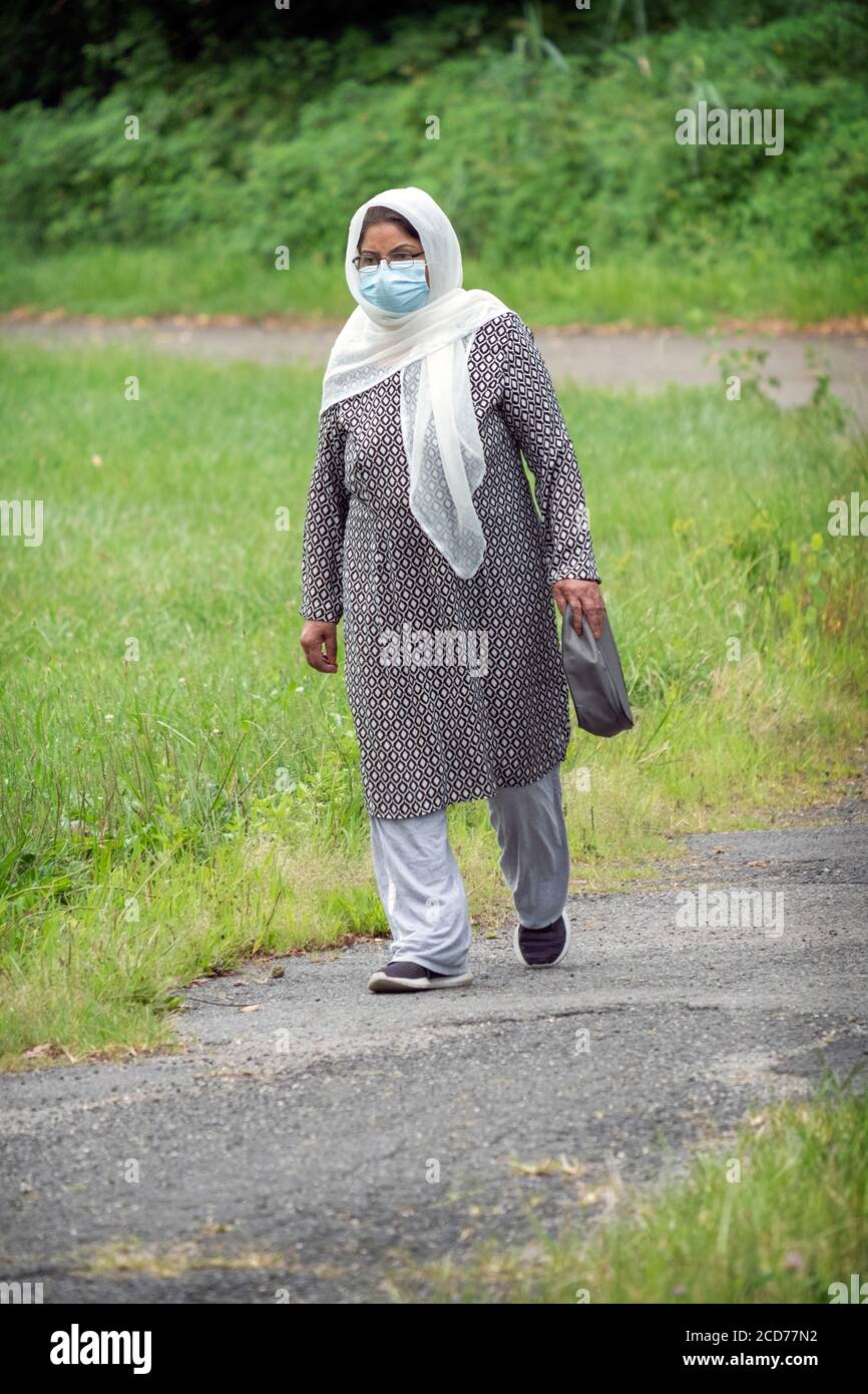 A Muslim American woman in traditional ethnic attire walks in a park in Queens, NYC while also wearing a surgical mask. Stock Photo