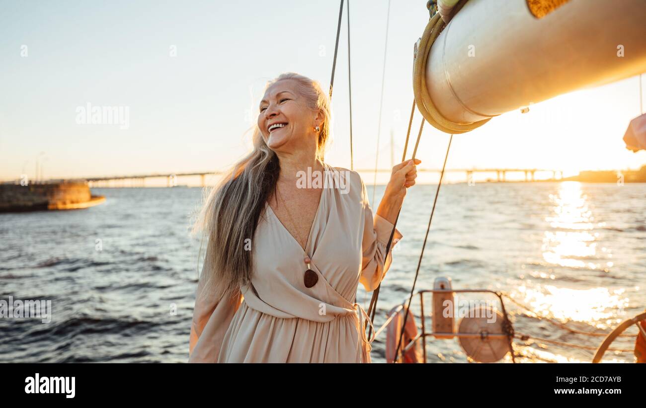 Happy mature woman standing on a yacht at sunset and looking away Stock Photo