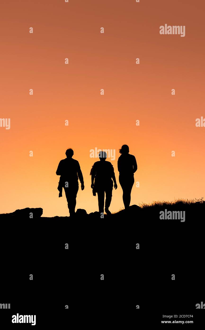 Three people walking on Pentire Point East silhouetted by an intense colourful sunset in Newquay in Cornwall. Stock Photo