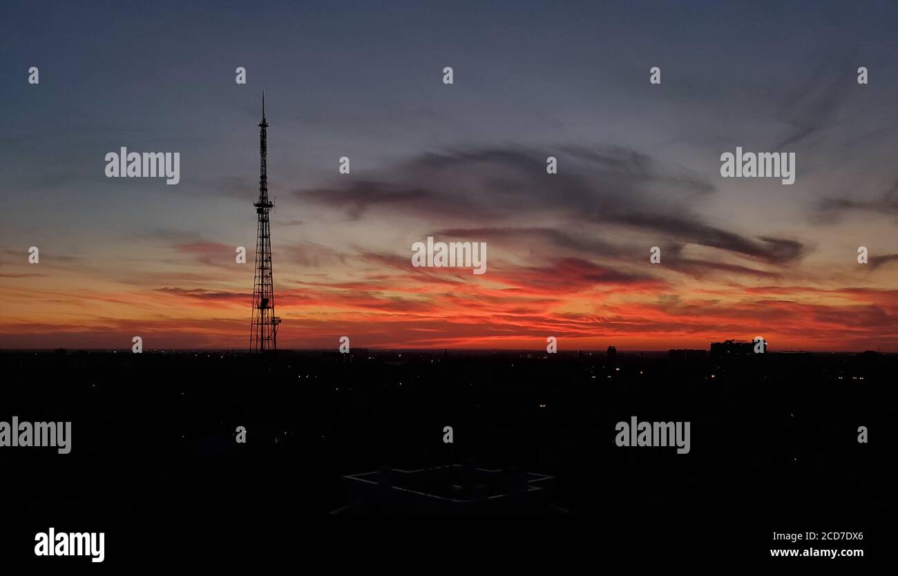 Blurry landscape of breathtaking sunset over urban skyline with communication tower silhouette. Purple and red clouds on dark evening sky background Stock Photo
