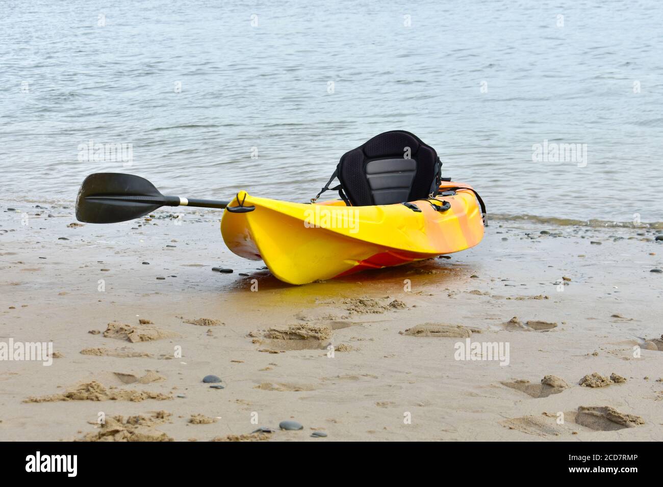 isolated bright yellow kayak with black canoe paddle and seat on the sand with sea lapping