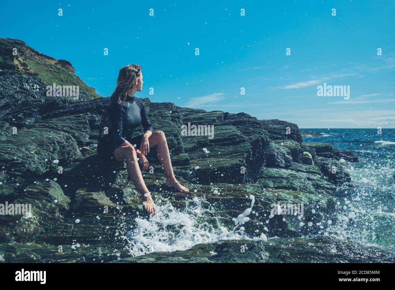 A young woman wearing a wetsuit is sitting on the rocks by the sea as ...