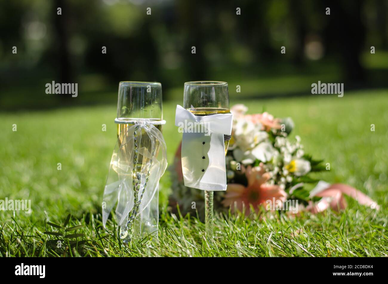 Two wedding glasses of champagne stand on the grass, against the background of a wedding bouquet. Sunny day. Close up Stock Photo