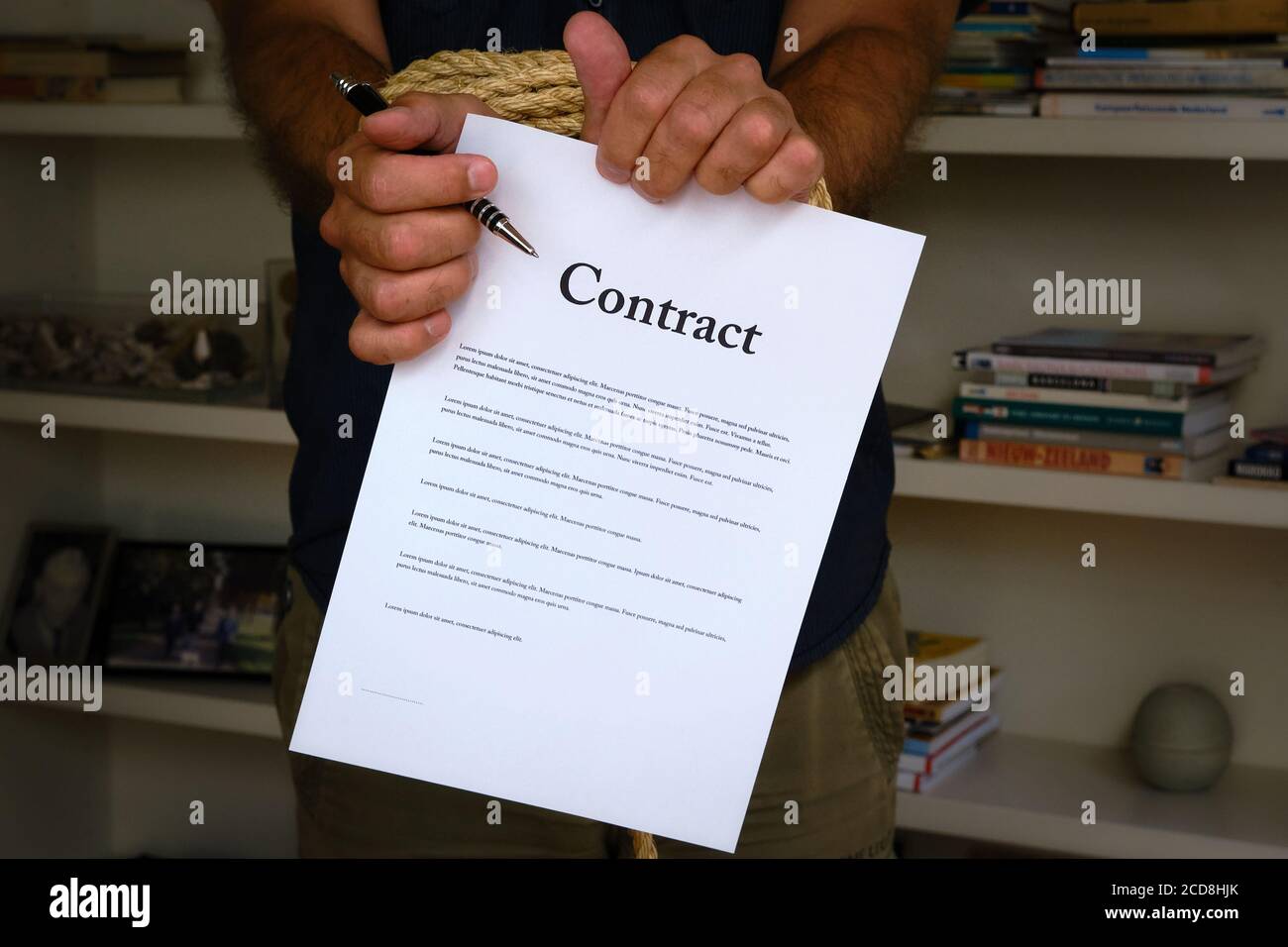 Tied hands holding pen and tearing contract paper in front of bookcase. Bound man hands rip paper contract, breach of contract. Unfair agreement Stock Photo