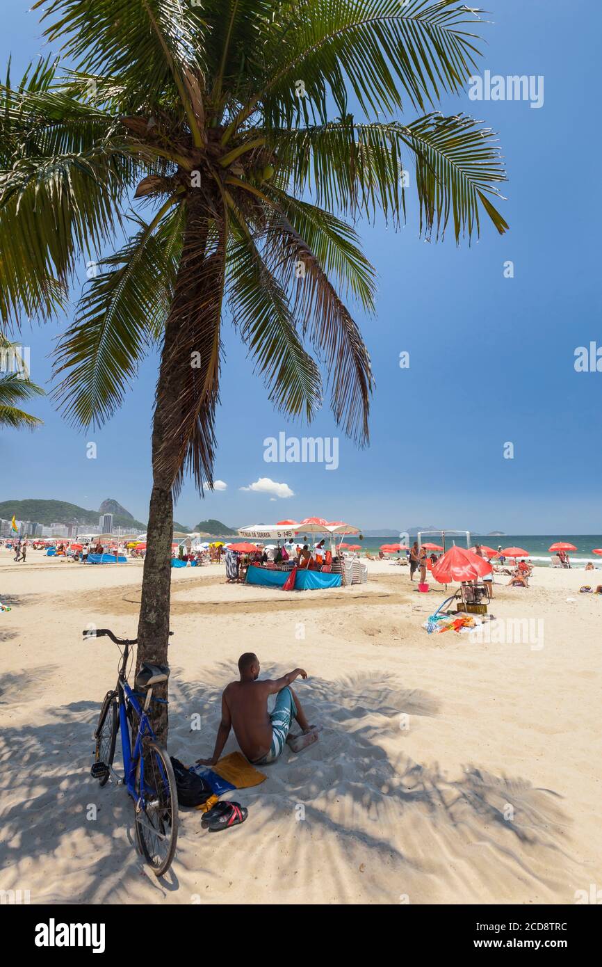 Brazil, Rio de Janeiro state, Rio de Janeiro city, Copacabana beach, Carioca landscapes between the mountain and the sea classified UNESCO World Heritage Site, man sitting on the sand, in a coconut tree shadow and looking the beach Stock Photo