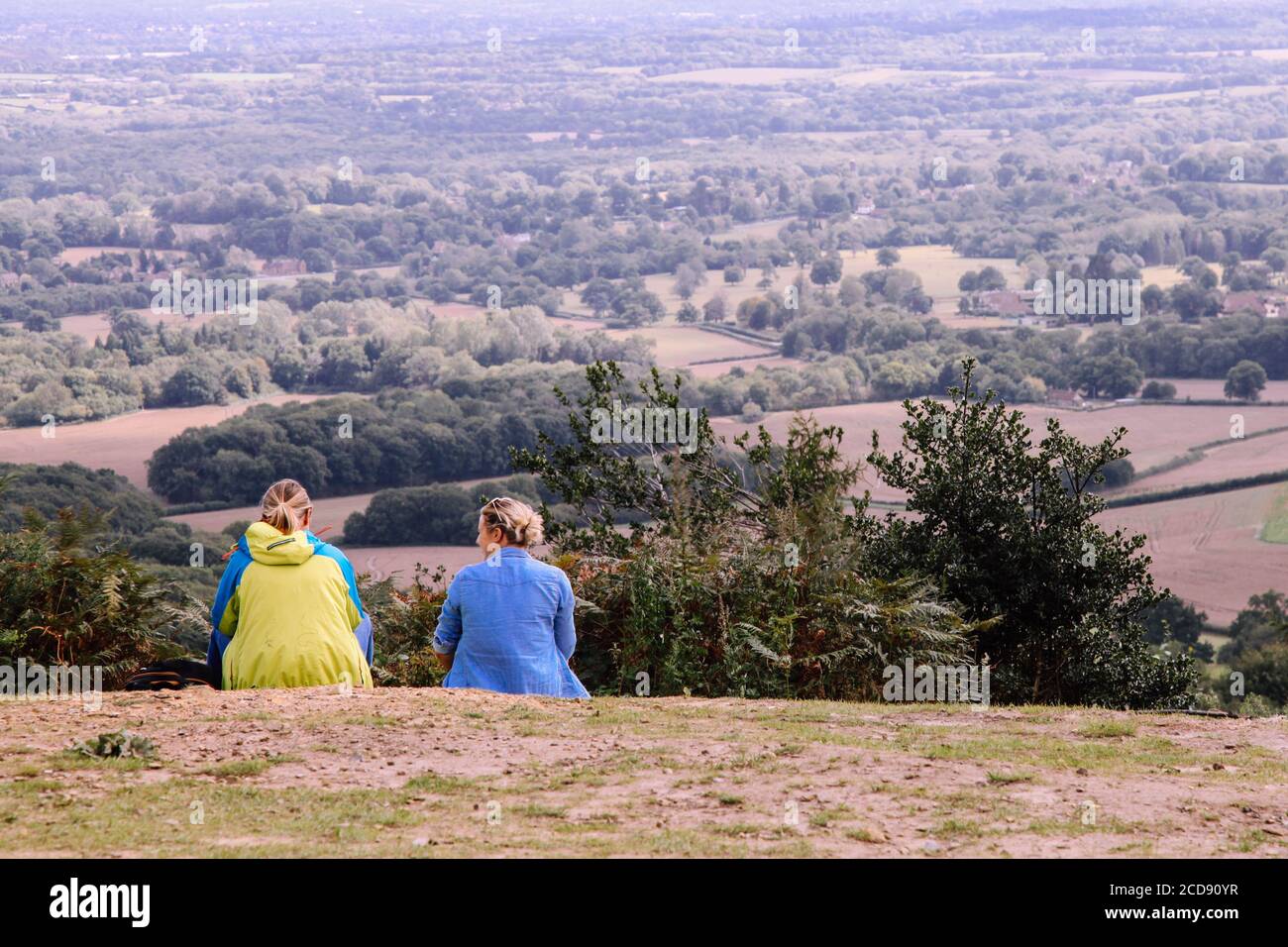 Two women sit on a hill talking and looking out at Surrey Hills countryside from behind, England, UK, August 2020 Stock Photo