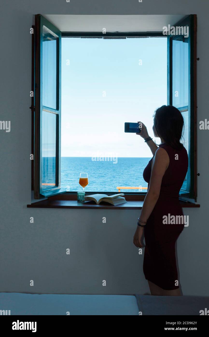 Spain, Canary Islands, La Palma, young and pretty woman taking a picture of the horizon from inside a hotel room, through a window, by the sea Stock Photo