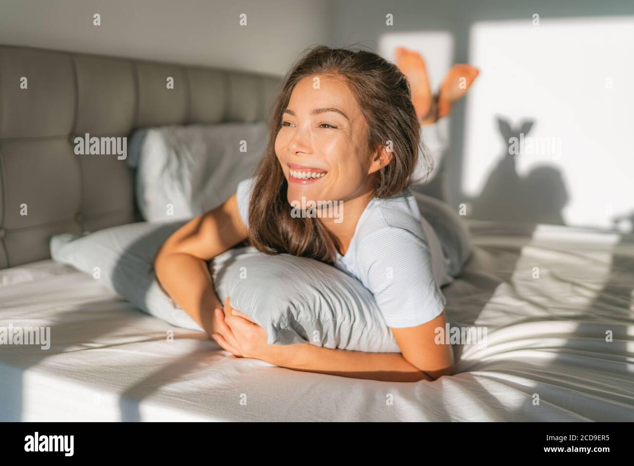 Bed woman waking up early morning happy enjoying sun on comfortable mattress and pillow. Asian girl relaxing in bedroom smiling. Natural beauty after Stock Photo