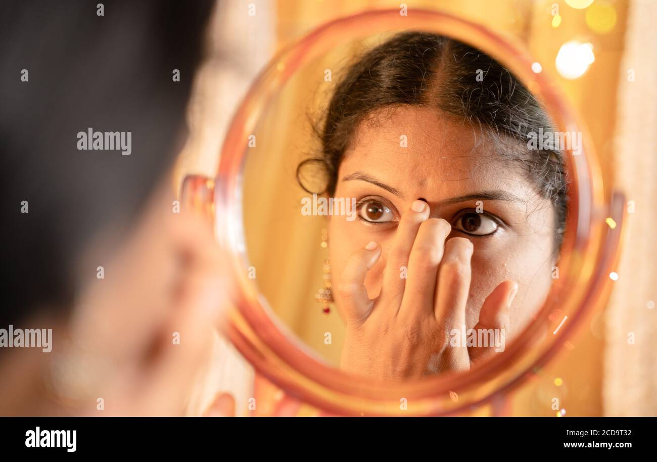 Indian married Woman applying Bindi, sindoor or decorative mark to forehead in front of mirror during festival celebrations with decoration lights as Stock Photo