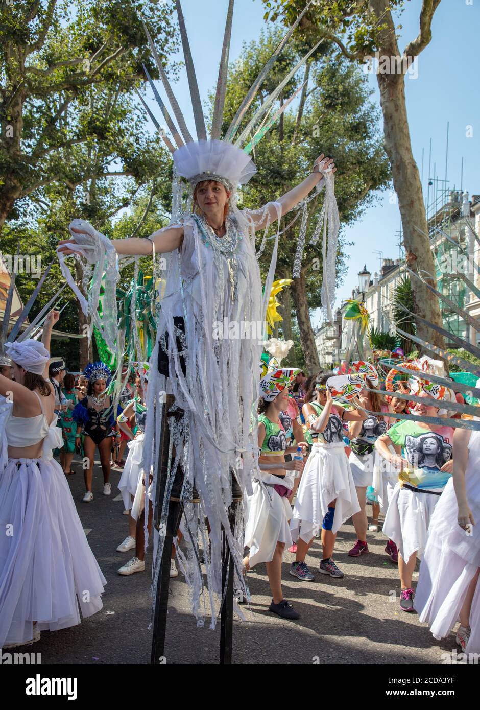 Woman on stilts, dressed in white, standing in front of dancers, taking