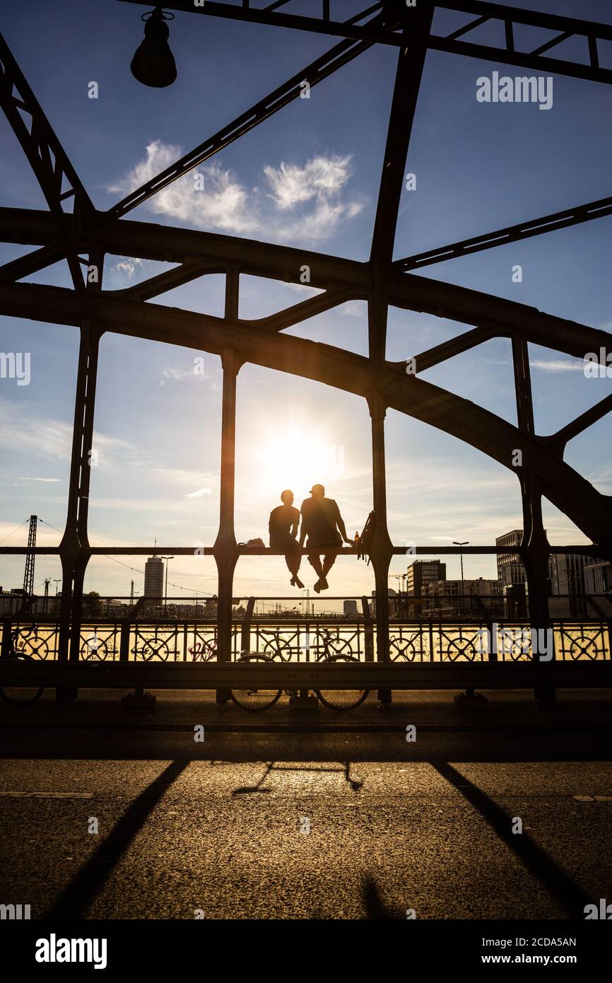 Unidentifiable backlit people enjoying the evening sun sitting on a steel bridge construction over railway tracks called Hackerbrücke. Munich, Germany Stock Photo