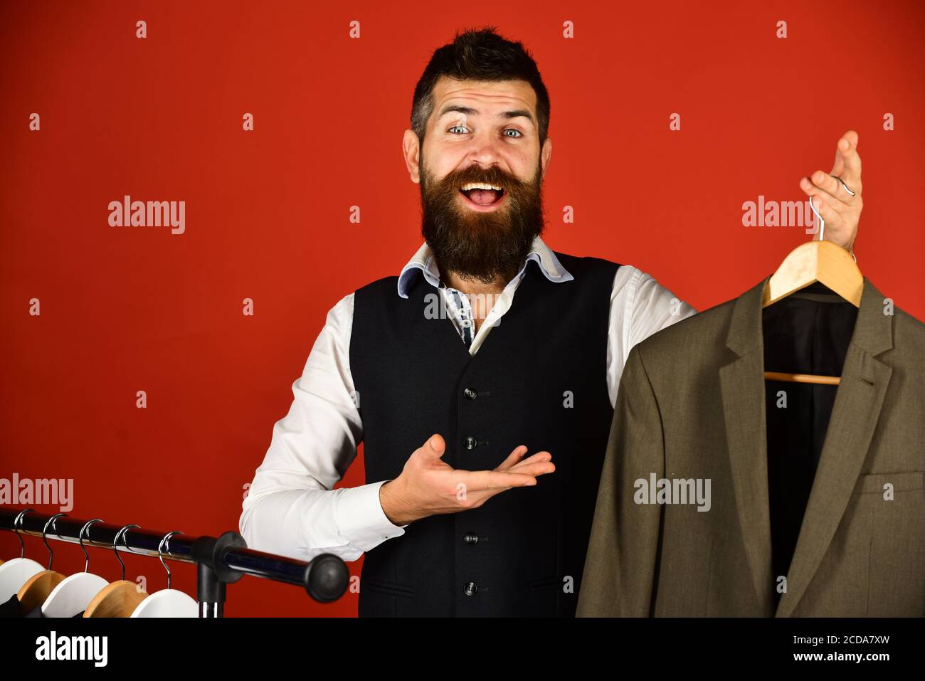 Handsome male customer by clothes rack holds brown jacket. Man with beard  and happy face points