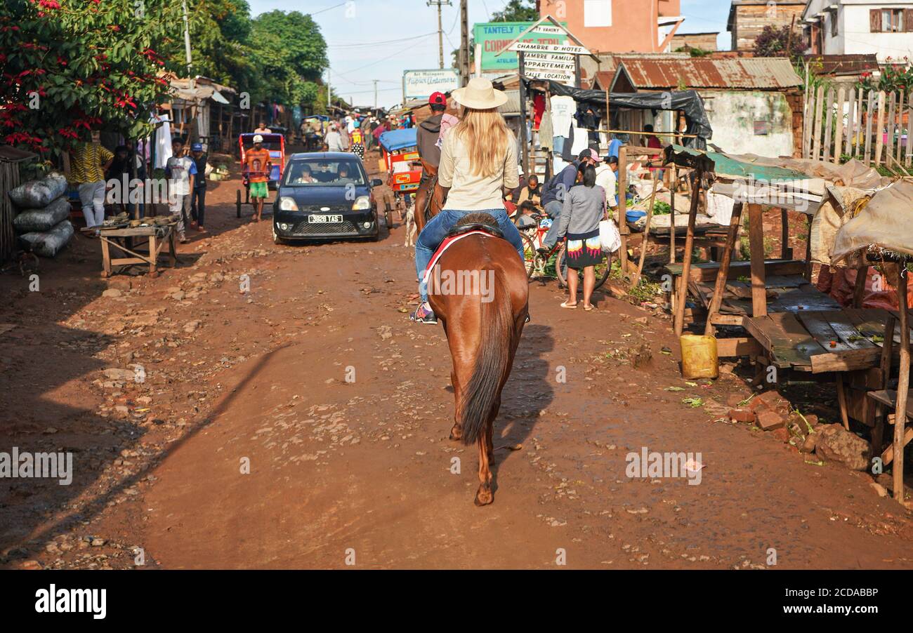 Antsirabe, Madagascar - April 26, 2019: Young woman in straw hat rides on her horse through morning market in Antsirabe, stalls on both sides, car com Stock Photo