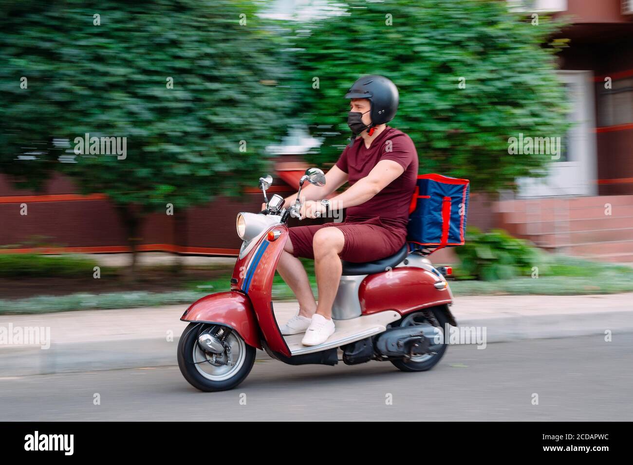 Delivery boy wearing red uniform on scooter with isothermal food case box driving fast. Express food delivery service from cafes and restaurants Stock Photo