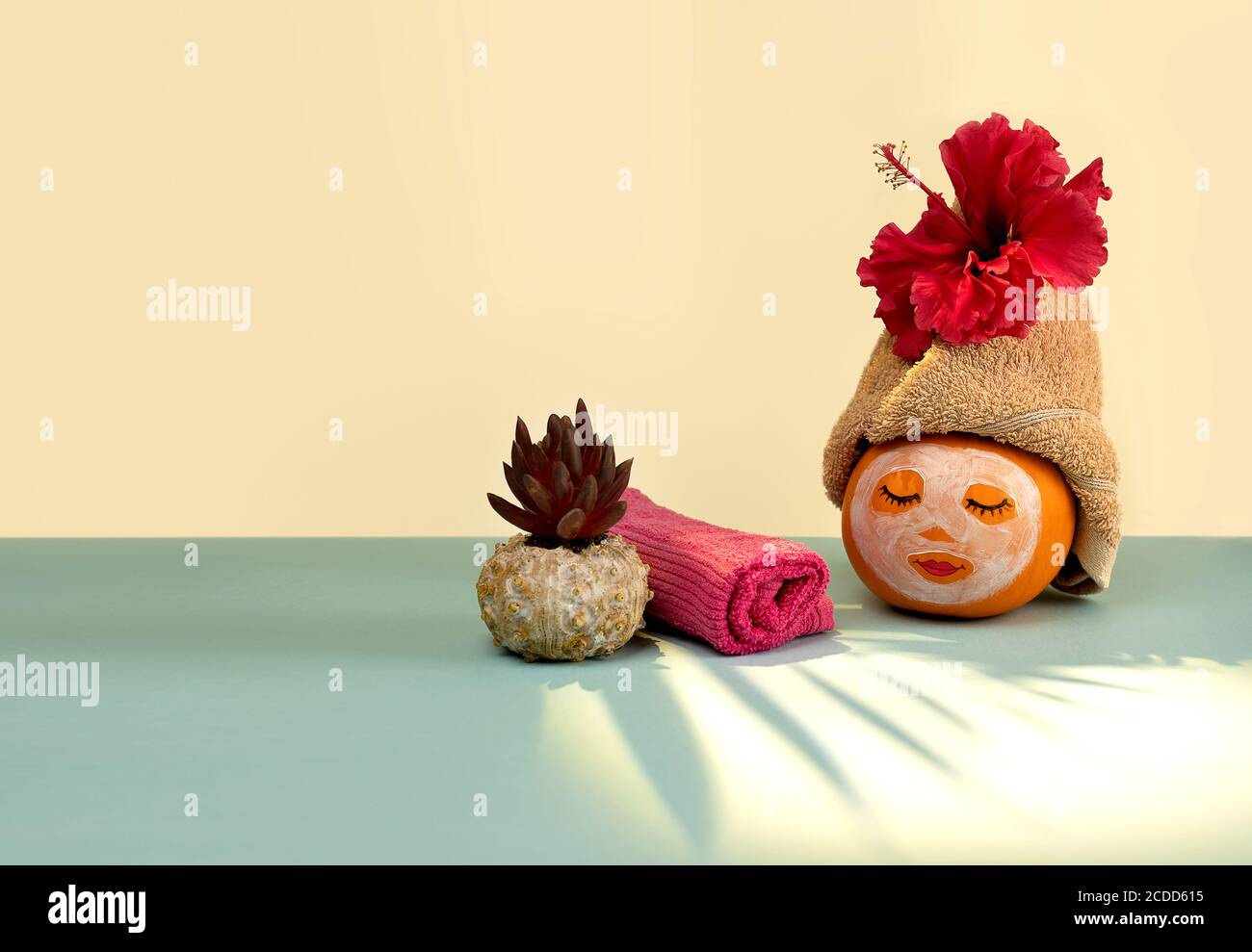 Pumpkin with painted face and facial mask, towel and flowers on pastel yellow and blue background with shadow from palm leaves. Spa concept, Helloween, Copy space. Stock Photo