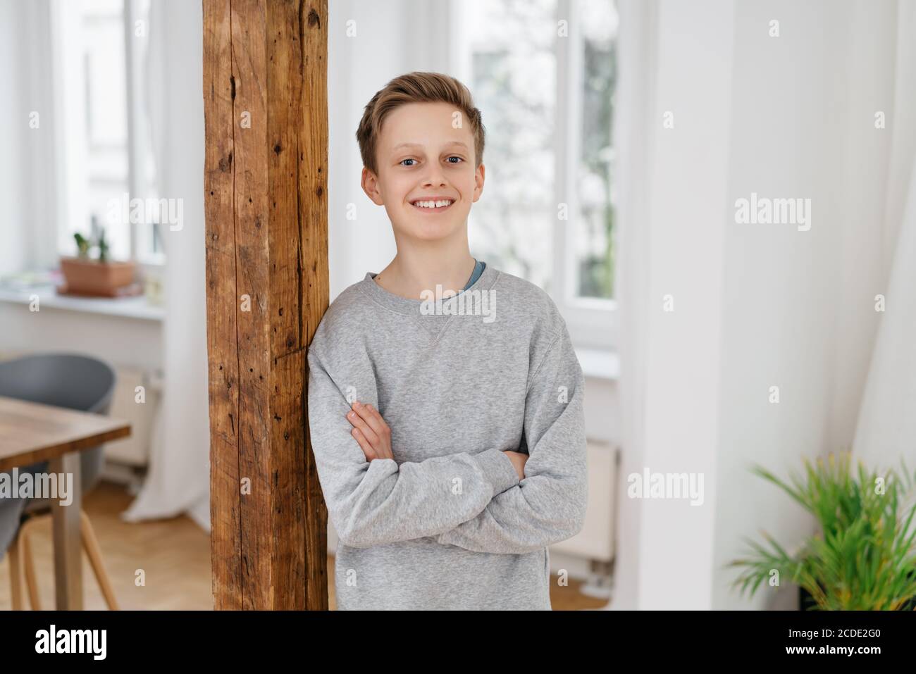 Self-assured young boy with a happy friendly smile standing leaning against a wooden interior pillar with folded arms in an apartment Stock Photo