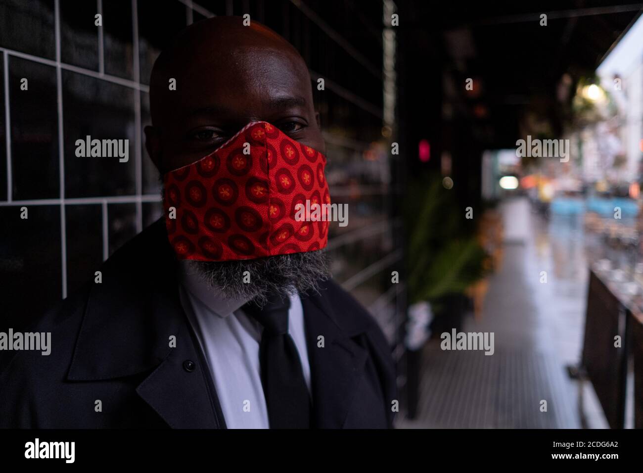 On a rainy night in Soho, security man 'H'  wears a bright red facial covering outside a business at a time when recently re-opened bars and restaurants are desperate for customer business during the coronavirus pandemic, on 27th August 2020, in London, England. Stock Photo