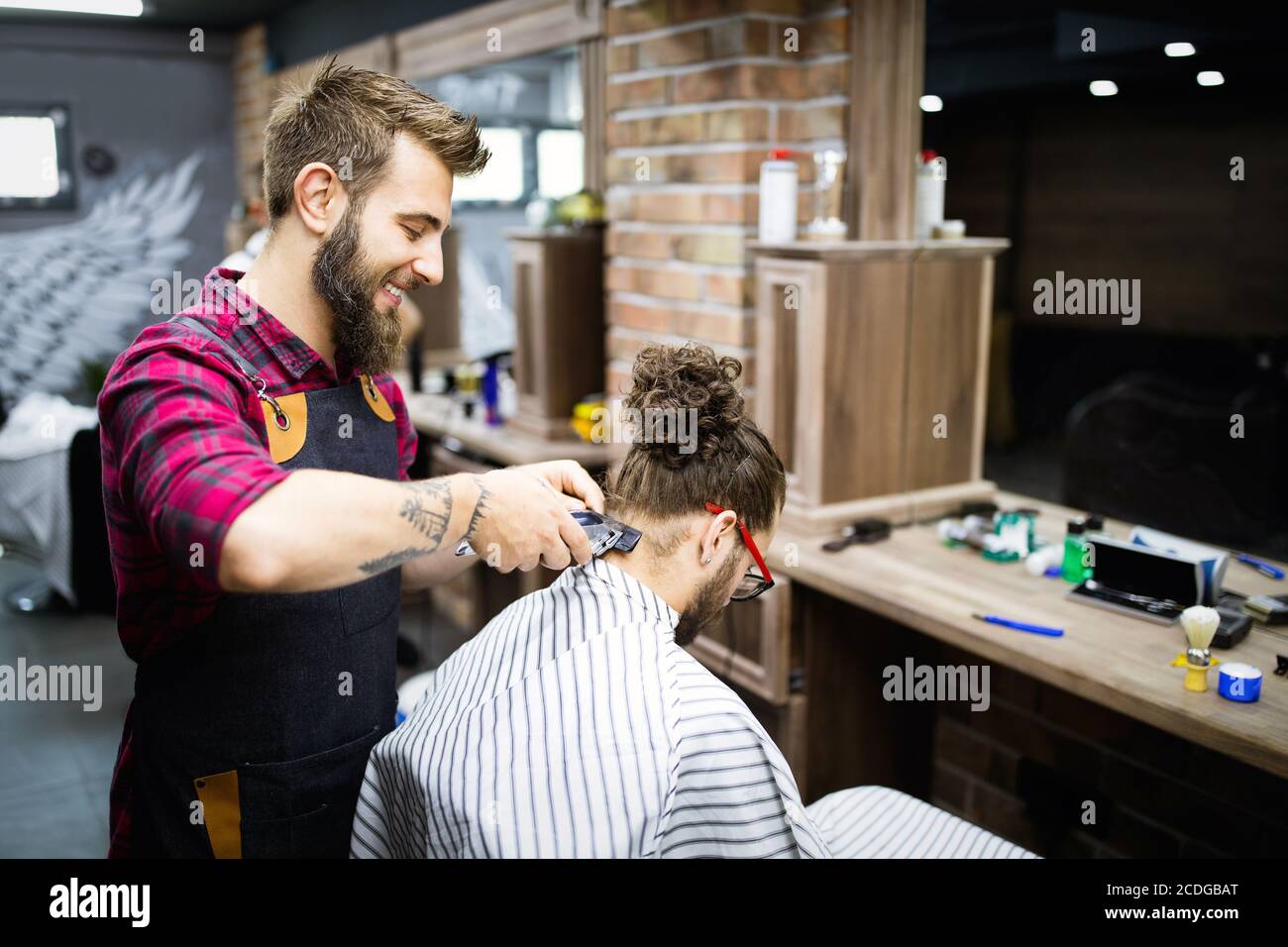 Young bearded man getting haircut by hairdresser with electric razor at barber shop Stock Photo