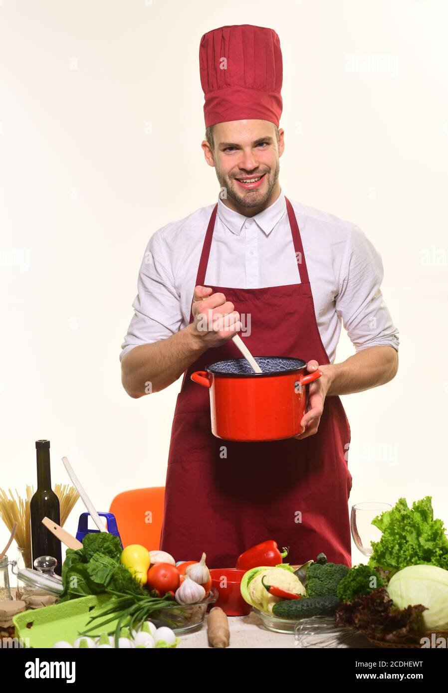 Man with beard isolated on white background. Cooking process concept. Chef  in burgundy uniform holds soup pot and ladle. Cook stands by kitchen table  with kitchenware and vegetables Stock Photo - Alamy