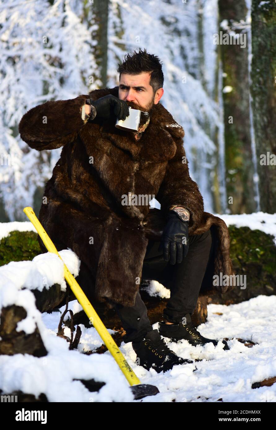 Macho with beard and mustache in forest. Guy on snowy nature park background. Man in fur coat drinks from metal flask near yellow axe. Brutal gamekeeper concept. Stock Photo