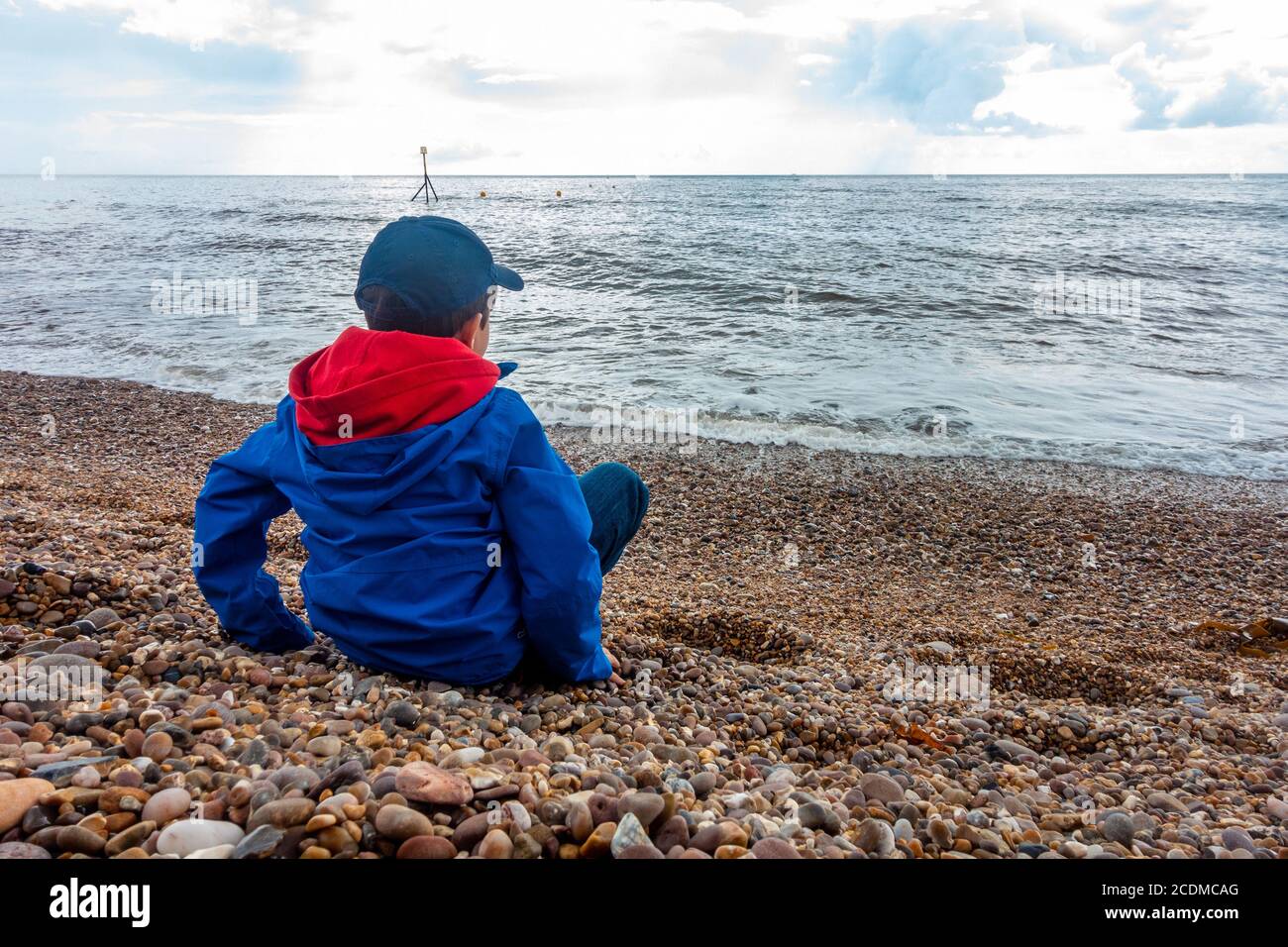 A young boy wearing a waterproof jacket sits on a quiet, pebbly beach at Sidmouth, Devon, UK and looks out to sea. Stock Photo