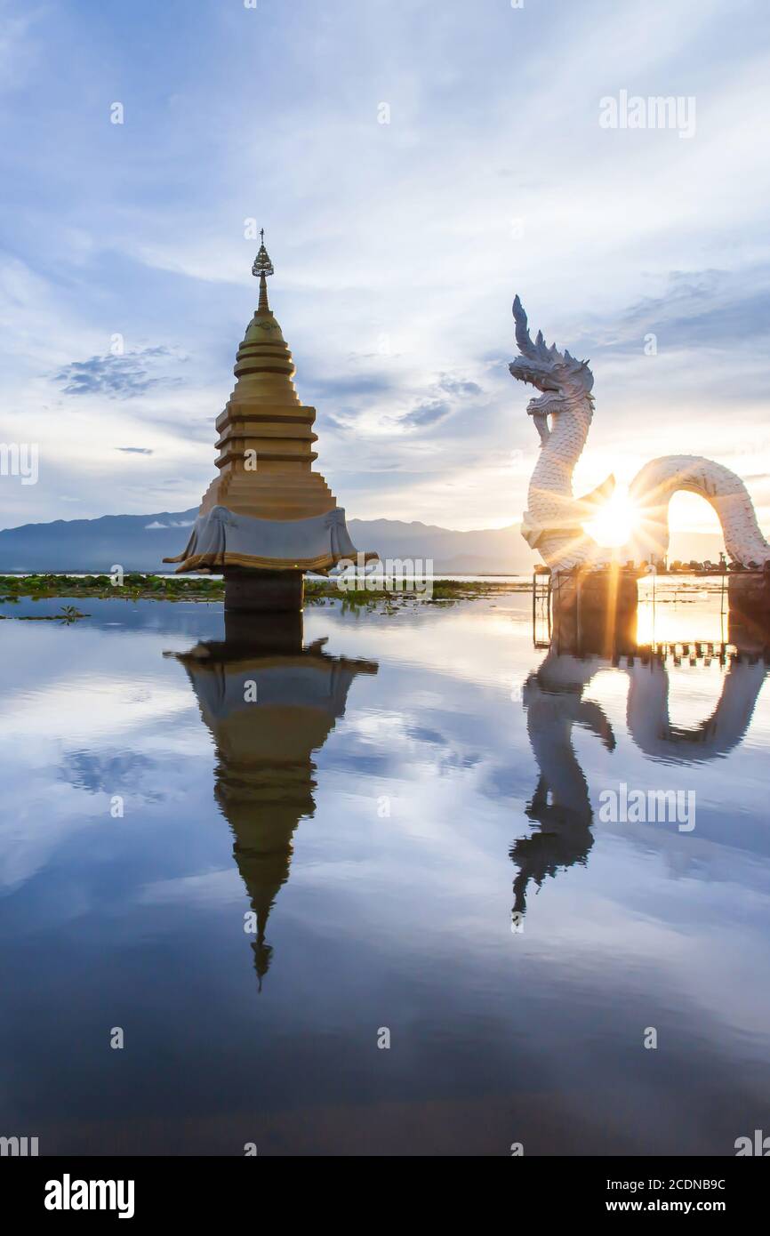 Naga statue and golden stupa on the lake at dusk, glowing sun setting through the Naga statue. Phayao Lake, Thailand. Stock Photo