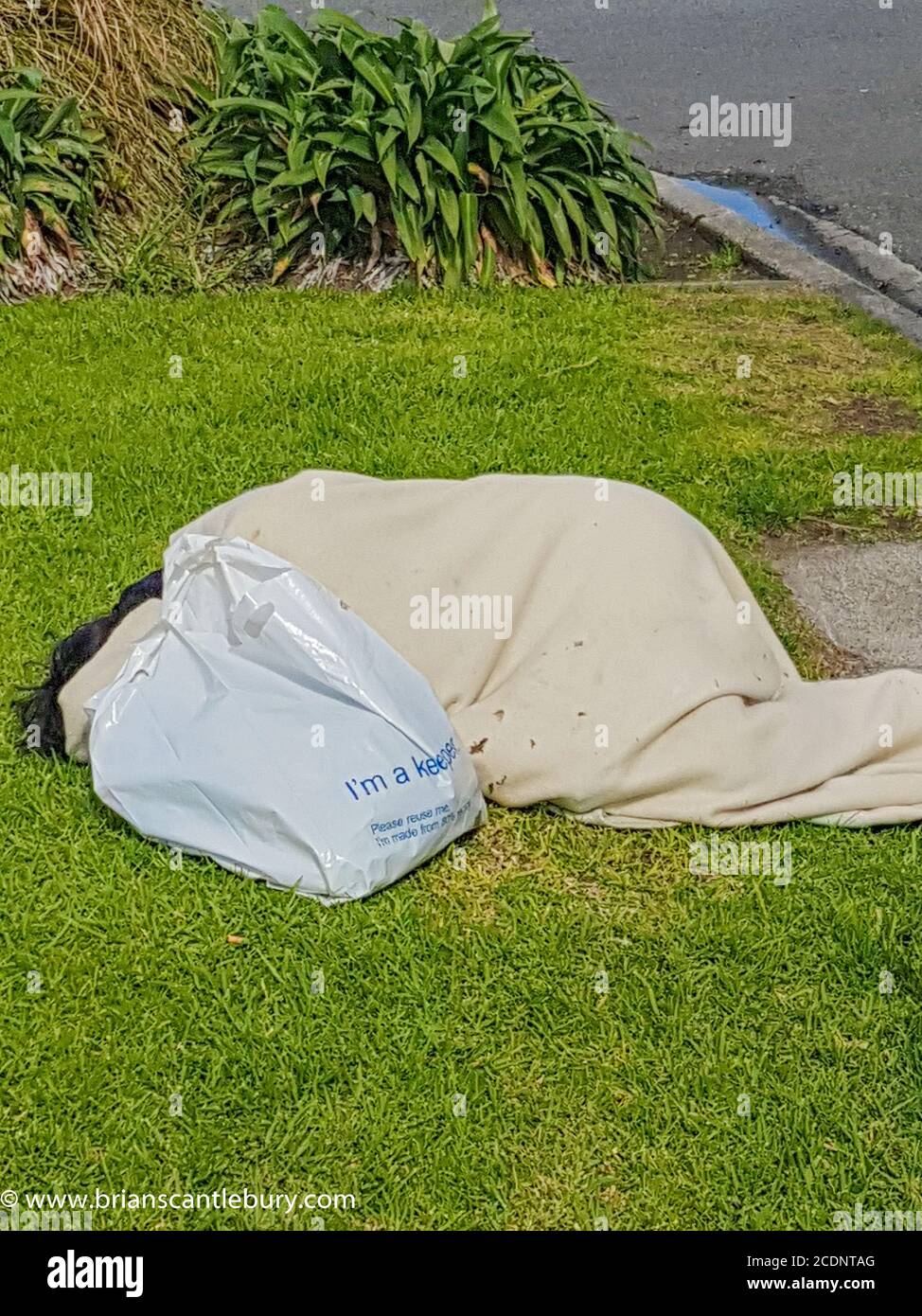 Tauranga New Zealand - August 29 2020; Homeless person asleep on grass in public area wrapped in cloth with plastic bag with words - I am a keeper. Stock Photo