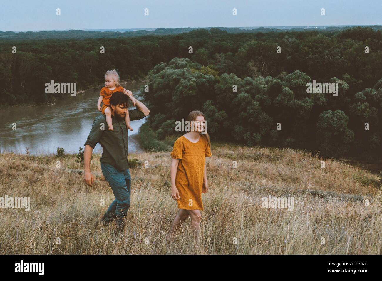 Family parents walking with child outdoor mother and father carrying baby daughter on shoulders healthy lifestyle rural nature river and forest view Stock Photo