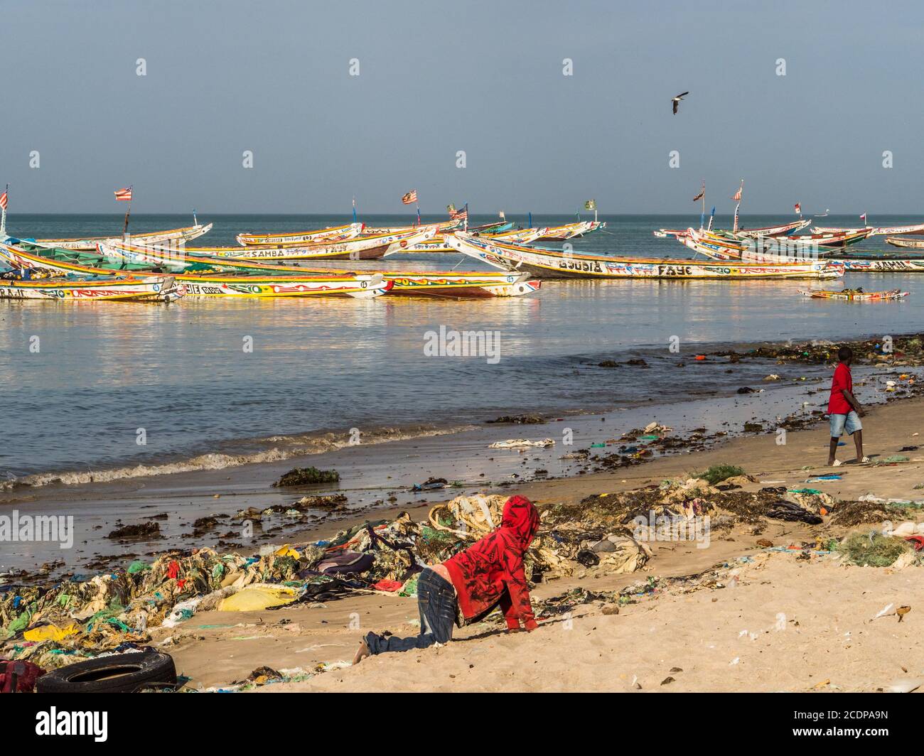 Senegal, Africa - January 26, 2019:  Chidren playing on  the beach with plenty of waste. Pollution concept. Colorful fisher boats in the background. S Stock Photo
